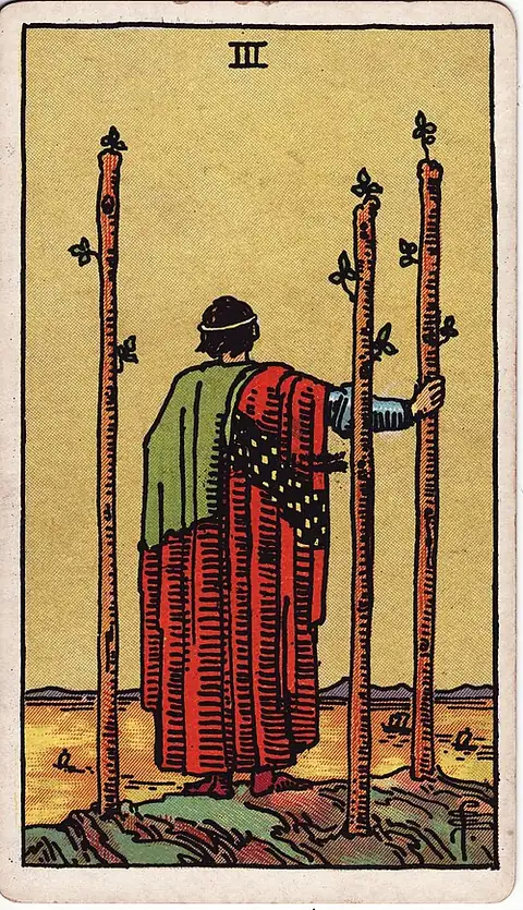 Three of Wands. A cloaked figure stands on a cliff among three planted staves, watching ships pass on the water below.