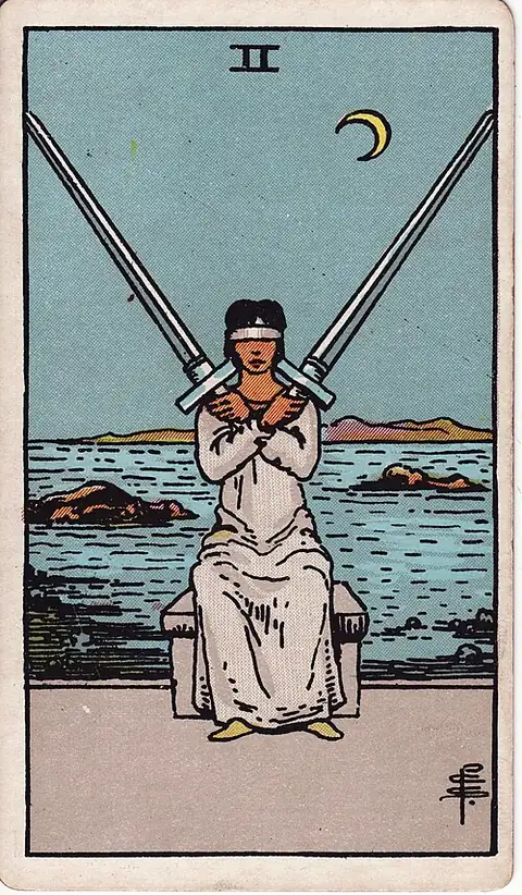 Two of Swords. A blindfolded woman sits with two swords crossed over her chest, a rocky sea and a crescent moon behind.